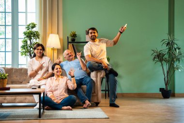 Sıcak ve terli hisseden Hintli aile, yaşlı ebeveynler ve genç yetişkin kız ve erkek çocuk modern oturma odasındaki koltukta oturuyorlar, kız klimayı uzaktan kumandayla açıyor, hepsi rahatlamış hissediyorlar.