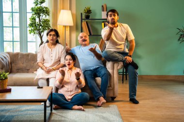 Kameraya şaşırmış, mutlu Hintli aile, yaşlı aileler ve genç yetişkin kız ve erkek çocuk modern oturma odasındaki koltukta oturmuş evdeki şaşırtıcı derecede komik bir şeye tepki gösteriyorlar.