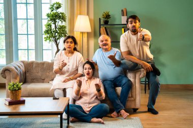 Kameraya şaşırmış, mutlu Hintli aile, yaşlı aileler ve genç yetişkin kız ve erkek çocuk modern oturma odasındaki koltukta oturmuş evdeki şaşırtıcı derecede komik bir şeye tepki gösteriyorlar.