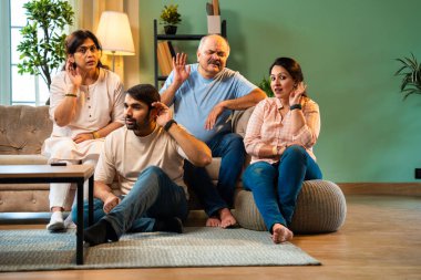 Kameraya bakarken kulakları, genç yetişkin bir oğlu ve kızı olan Hintli bir aile rahat modern oturma odasında oturup dedikodu ya da gizli şeyler dinliyorlar.