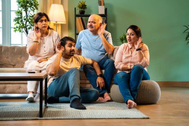 Kameraya bakarken kulakları, genç yetişkin bir oğlu ve kızı olan Hintli bir aile rahat modern oturma odasında oturup dedikodu ya da gizli şeyler dinliyorlar.