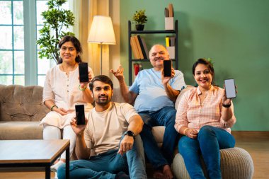 Boş bir telefon ekranı, dört çocuklu Hintli aile, evde rahat bir koltukta oturmuş gülümseyen ve kameraya uygulamayı veya servisi destekleyen yetişkin aile ve çocuklar.
