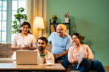 İnternetten alışveriş yaparken kanepede oturan, kıdemli ebeveynleri ve küçük yetişkin çocukları olan neşeli Hintli aile, evlerinde dizüstü bilgisayar ve kredi kartı kullanarak dijital ödeme yaparken kaliteli vakit geçirmenin keyfini çıkarıyor.