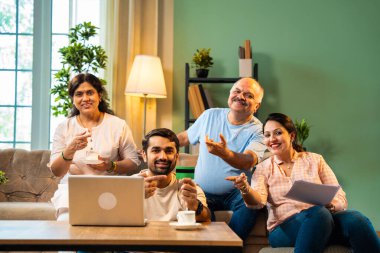 İnternetten alışveriş yaparken kanepede oturan, kıdemli ebeveynleri ve küçük yetişkin çocukları olan neşeli Hintli aile, evlerinde dizüstü bilgisayar ve kredi kartı kullanarak dijital ödeme yaparken kaliteli vakit geçirmenin keyfini çıkarıyor.