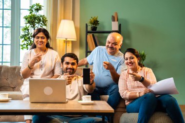 Boş bir telefon ekranı, dört çocuklu Hintli aile, evde rahat bir koltukta oturmuş gülümseyen ve kameraya uygulamayı veya servisi destekleyen yetişkin aile ve çocuklar.