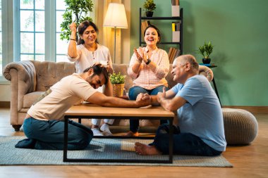 Hindistan 'ın kıdemli babasıyla genç yetişkin oğlu arasında bilek güreşi eğlencesi. Kıdemli eş ve genç kız gülüyor ve tezahürat yapıyorlar. Evde arkadaşça eğleniyorlar.