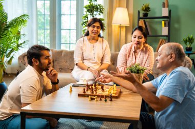 Evde kanepede satranç oynarken, dört çocuklu Hintli aile, yaşlı ebeveynler ve genç yetişkin çocuklar dahil modern konforlu oturma odasında kaliteli zaman geçirmenin keyfini çıkarıyorlar, akıllı masa oyunlarıyla kaynaşıyorlar.