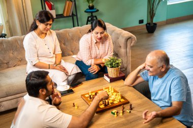 Evde kanepede satranç oynarken, dört çocuklu Hintli aile, yaşlı ebeveynler ve genç yetişkin çocuklar dahil modern konforlu oturma odasında kaliteli zaman geçirmenin keyfini çıkarıyorlar, akıllı masa oyunlarıyla kaynaşıyorlar.
