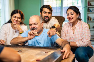 Birlikte carom ya da carrom masa üstü oyunu oynayarak, dört kişilik Hintli aile, yaşlı ebeveynler ve genç yetişkin bir çocuk ve kız evdeki boş zamanlarında rahat oturma odasında eğleniyorlar.