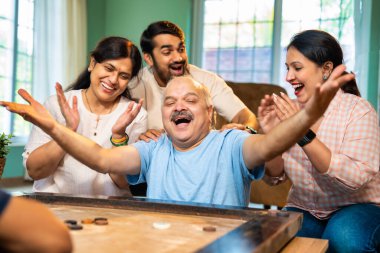 Birlikte carom ya da carrom masa üstü oyunu oynayarak, dört kişilik Hintli aile, yaşlı ebeveynler ve genç yetişkin bir çocuk ve kız evdeki boş zamanlarında rahat oturma odasında eğleniyorlar.