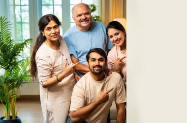 Boş bir tahtanın arkasından gelen ve gülümseyen dört kişilik Hintli aile, kıdemli ebeveynler ve genç yetişkin kız ve erkek çocuk reklam mesajları ya da kapalı alandaki beyaz fotokopi alanından yapılan duyurular.