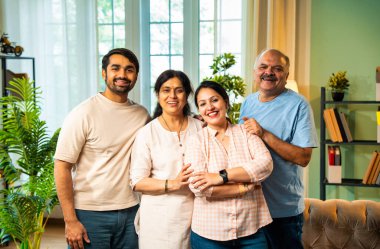 Kameraya bakıp gülümseyen, dört çocuklu Hintli aile, kıdemli ebeveynler ve genç yetişkin kız ve erkek çocuklar modern oturma odasında birlikte poz veriyorlar. Mutlu ve birlik olmuş görünüyorlar.
