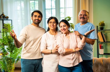 Kameraya bakıp gülümseyen, dört çocuklu Hintli aile, kıdemli ebeveynler ve genç yetişkin kız ve erkek çocuklar modern oturma odasında birlikte poz veriyorlar. Mutlu ve birlik olmuş görünüyorlar.