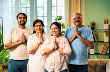 namaskar duruşu yapan ve kameraya gülümseyen dört kişilik Hintli aile, yaşlı ebeveynler ve genç yetişkin kız ve erkek çocukları, samimi, modern ev dekorasyonlarıyla selamlıyorlar.