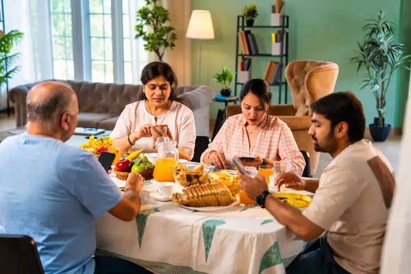Birlikte kahvaltı yapıyorlar, Hintli üst düzey ebeveynler ve genç yetişkin kız ve erkek çocuklar rahat bir evde, modern yemek masasında sessizce oturuyorlar, akıllı telefonlar kullanıyorlar, sabah yemeği sırasında dijital olarak dikkatleri dağılıyorlar.