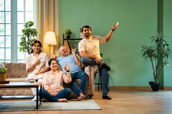 Sıcak ve terli hisseden Hintli aile, yaşlı ebeveynler ve genç yetişkin kız ve erkek çocuk modern oturma odasındaki koltukta oturuyorlar, kız klimayı uzaktan kumandayla açıyor, hepsi rahatlamış hissediyorlar.