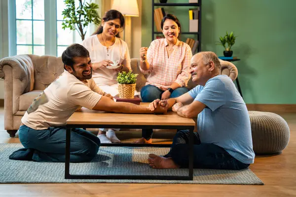 Hindistan 'ın kıdemli babasıyla genç yetişkin oğlu arasında bilek güreşi eğlencesi. Kıdemli eş ve genç kız gülüyor ve tezahürat yapıyorlar. Evde arkadaşça eğleniyorlar.