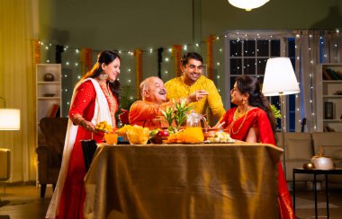 Laddu ya da laddu yiyip gülümseyen dört çocuklu Hintli aile, yaşlı ebeveynleriyle Diwali festivalini evde kutluyor, dekore edilmiş yemek masasında oturmuş neşeyle kameraya bakıyor.