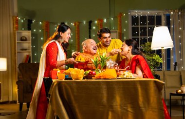 Laddu ya da laddu yiyip gülümseyen dört çocuklu Hintli aile, yaşlı ebeveynleriyle Diwali festivalini evde kutluyor, dekore edilmiş yemek masasında oturmuş neşeyle kameraya bakıyor.