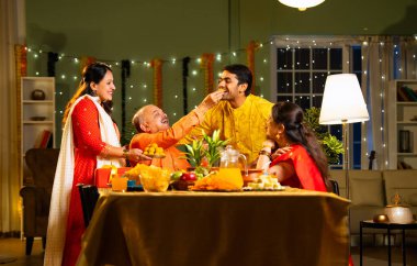 Laddu ya da laddu yiyip gülümseyen dört çocuklu Hintli aile, yaşlı ebeveynleriyle Diwali festivalini evde kutluyor, dekore edilmiş yemek masasında oturmuş neşeyle kameraya bakıyor.