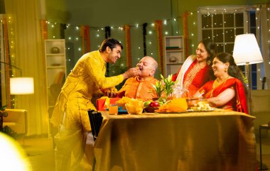 Laddu ya da laddu yiyip gülümseyen dört çocuklu Hintli aile, yaşlı ebeveynleriyle Diwali festivalini evde kutluyor, dekore edilmiş yemek masasında oturmuş neşeyle kameraya bakıyor.