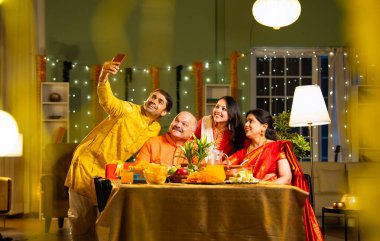 Yemek masasında selfie çeken dört kişilik Hintli aile, kıdemli ebeveynleri, yetişkin oğulları ve kızları ile Diwali festivalini içeride dekorasyonlar, tatlılar ve gülücüklerle kutluyor.
