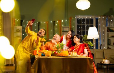 Yemek masasında selfie çeken dört kişilik Hintli aile, kıdemli ebeveynleri, yetişkin oğulları ve kızları ile Diwali festivalini içeride dekorasyonlar, tatlılar ve gülücüklerle kutluyor.