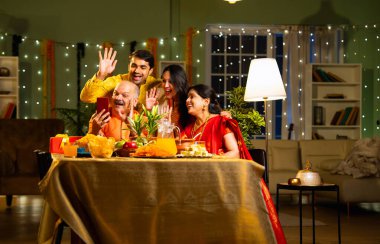 Video konferansında Hint ailesi, Hindu festivalini evde kutlayan yaşlı ebeveynler ve genç yetişkin çocuklar, evde dekore edilmiş yemek masasındaki akrabalarına gülümsüyor ve onlarla konuşuyor.