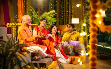 Ladus 'u kucaklayan ve birlikte gülümseyen Hintli aile, genç bir kızla birlikte, sari giymiş yaşlı bir anne, geleneksel giysili baba ve koca Diwali' yi kapalı mekanda kutluyor, çiçek rangoli ve kahkahalarla.