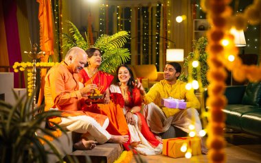 Ladus 'u kucaklayan ve birlikte gülümseyen Hintli aile, genç bir kızla birlikte, sari giymiş yaşlı bir anne, geleneksel giysili baba ve koca Diwali' yi kapalı mekanda kutluyor, çiçek rangoli ve kahkahalarla.