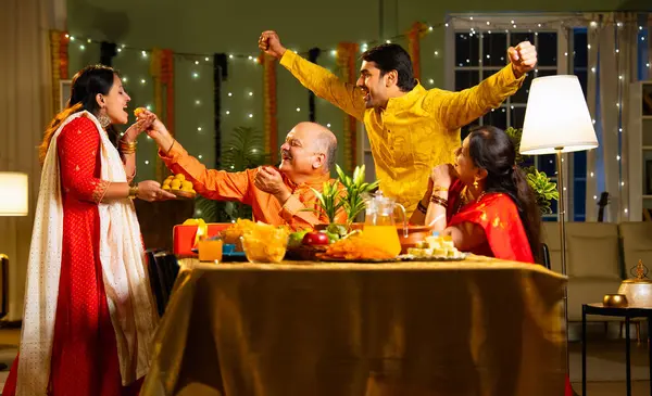 Laddu ya da laddu yiyip gülümseyen dört çocuklu Hintli aile, yaşlı ebeveynleriyle Diwali festivalini evde kutluyor, dekore edilmiş yemek masasında oturmuş neşeyle kameraya bakıyor.