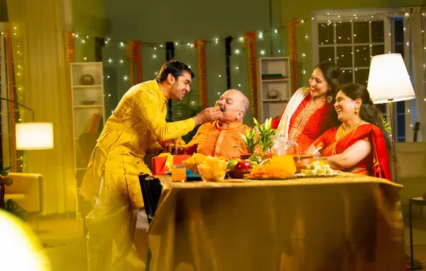 Laddu ya da laddu yiyip gülümseyen dört çocuklu Hintli aile, yaşlı ebeveynleriyle Diwali festivalini evde kutluyor, dekore edilmiş yemek masasında oturmuş neşeyle kameraya bakıyor.