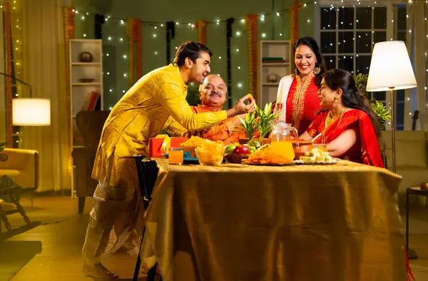 Laddu ya da laddu yiyip gülümseyen dört çocuklu Hintli aile, yaşlı ebeveynleriyle Diwali festivalini evde kutluyor, dekore edilmiş yemek masasında oturmuş neşeyle kameraya bakıyor.