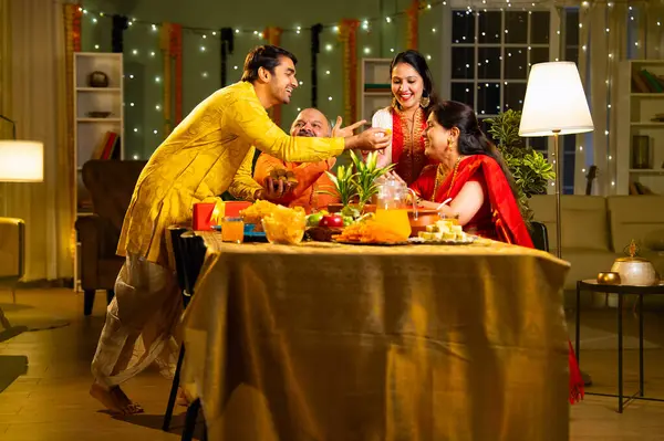 Laddu ya da laddu yiyip gülümseyen dört çocuklu Hintli aile, yaşlı ebeveynleriyle Diwali festivalini evde kutluyor, dekore edilmiş yemek masasında oturmuş neşeyle kameraya bakıyor.
