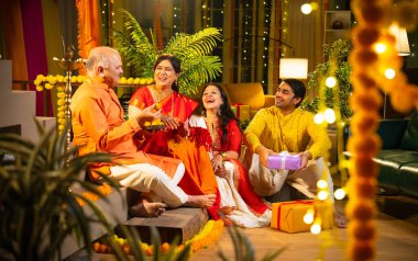 Ladus 'u kucaklayan ve birlikte gülümseyen Hintli aile, genç bir kızla birlikte, sari giymiş yaşlı bir anne, geleneksel giysili baba ve koca Diwali' yi kapalı mekanda kutluyor, çiçek rangoli ve kahkahalarla.