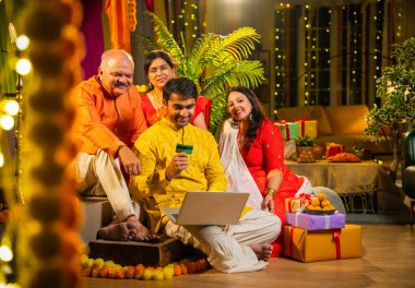 İnternetten bilgisayar ve kredi kartı kullanarak alışveriş yapan, karısı olan Hintli genç adam, geleneksel kıdemli ebeveynler evde Diwali gecesini kutluyorlar, para, çiçek ve ev dekorasyonlarıyla birlikte oturuyorlar.
