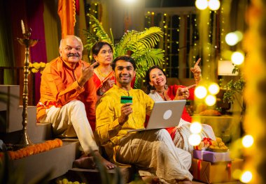 İnternetten bilgisayar ve kredi kartı kullanarak alışveriş yapan, karısı olan Hintli genç adam, geleneksel kıdemli ebeveynler evde Diwali gecesini kutluyorlar, para, çiçek ve ev dekorasyonlarıyla birlikte oturuyorlar.