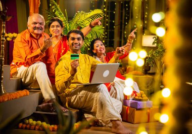 İnternetten bilgisayar ve kredi kartı kullanarak alışveriş yapan, karısı olan Hintli genç adam, geleneksel kıdemli ebeveynler evde Diwali gecesini kutluyorlar, para, çiçek ve ev dekorasyonlarıyla birlikte oturuyorlar.