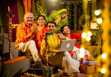 İnternetten bilgisayar ve kredi kartı kullanarak alışveriş yapan, karısı olan Hintli genç adam, geleneksel kıdemli ebeveynler evde Diwali gecesini kutluyorlar, para, çiçek ve ev dekorasyonlarıyla birlikte oturuyorlar.
