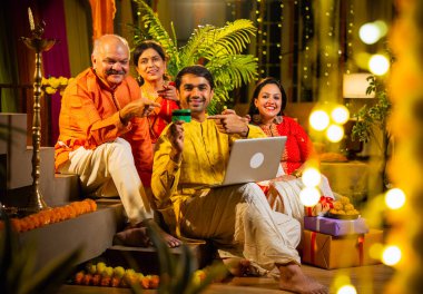 İnternetten bilgisayar ve kredi kartı kullanarak alışveriş yapan, karısı olan Hintli genç adam, geleneksel kıdemli ebeveynler evde Diwali gecesini kutluyorlar, para, çiçek ve ev dekorasyonlarıyla birlikte oturuyorlar.