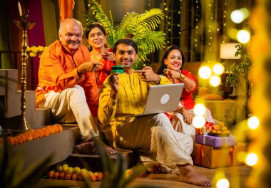 İnternetten bilgisayar ve kredi kartı kullanarak alışveriş yapan, karısı olan Hintli genç adam, geleneksel kıdemli ebeveynler evde Diwali gecesini kutluyorlar, para, çiçek ve ev dekorasyonlarıyla birlikte oturuyorlar.