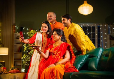 Diwali kutlaması sırasında selfie çeken, kıdemli ebeveynleri ve yetişkin çocukları olan Hintli aile akşam şenliklerinde birlikte oturan, ışıklarla süslenmiş, rahat koltukta mutlu bir şekilde gülümseyen.
