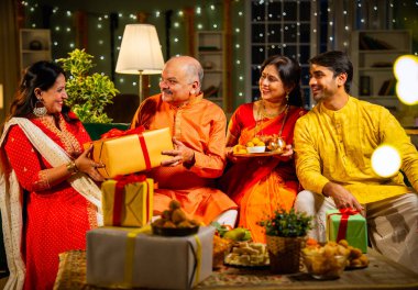 Yaşlı babaya Diwali hediyesi vermek, geleneksel giyimli Hintli kız festivalini evde kocası, annesi, laddu tabağı ve puja thali ile birlikte dekore edilmiş bir evde kanepede oturarak kutluyor.