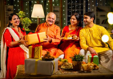 Yaşlı babaya Diwali hediyesi vermek, geleneksel giyimli Hintli kız festivalini evde kocası, annesi, laddu tabağı ve puja thali ile birlikte dekore edilmiş bir evde kanepede oturarak kutluyor.