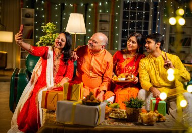 Diwali kutlaması sırasında selfie çeken, kıdemli ebeveynleri ve yetişkin çocukları olan Hintli aile akşam şenliklerinde birlikte oturan, ışıklarla süslenmiş, rahat koltukta mutlu bir şekilde gülümseyen.