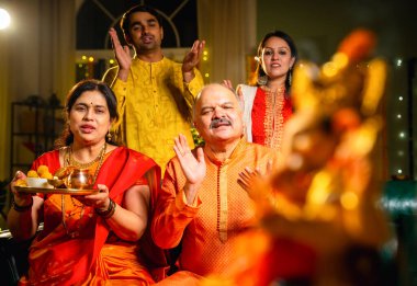 Ganesh Chaturthi sırasındaki Ganesha putuna odaklanın. Hintli aile, büyük anne-baba, oğul, gelin laddu tabağı ve puja thali ile süslenmiş festival evinde dua edin.