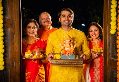 Ganesh Chaturthi sırasındaki Ganesha 'nın kil heykelini Hintli bir aile, yaşlı bir aile ve genç bir çift karşılıyor Ganpati' yi laddu ve puja thali ile dekore edilmiş evin girişinde ibadet için eve getiriyorlar.