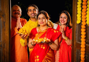 Puja thali ve laddu tabağıyla girişte duran, Hintli ailelere gülümseyen yaşlı aileler ve geleneksel şenlik elbisesi giyen genç çiftler süslü evde Hindu kutlamaları sırasında karşılanıyorlar.