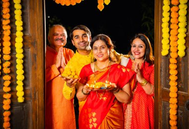 Puja thali ve laddu tabağıyla girişte duran, Hintli ailelere gülümseyen yaşlı aileler ve geleneksel şenlik elbisesi giyen genç çiftler süslü evde Hindu kutlamaları sırasında karşılanıyorlar.