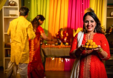 Ganesh Chaturthi kutlaması için davet edilen, geleneksel kıyafetli Hintli genç kız elinde laddu thali ile kameraya gülümsüyor, aile evi neşeyle hazırlarken neşeli puja 'ya katılmak için sıcak jestler yapıyor.